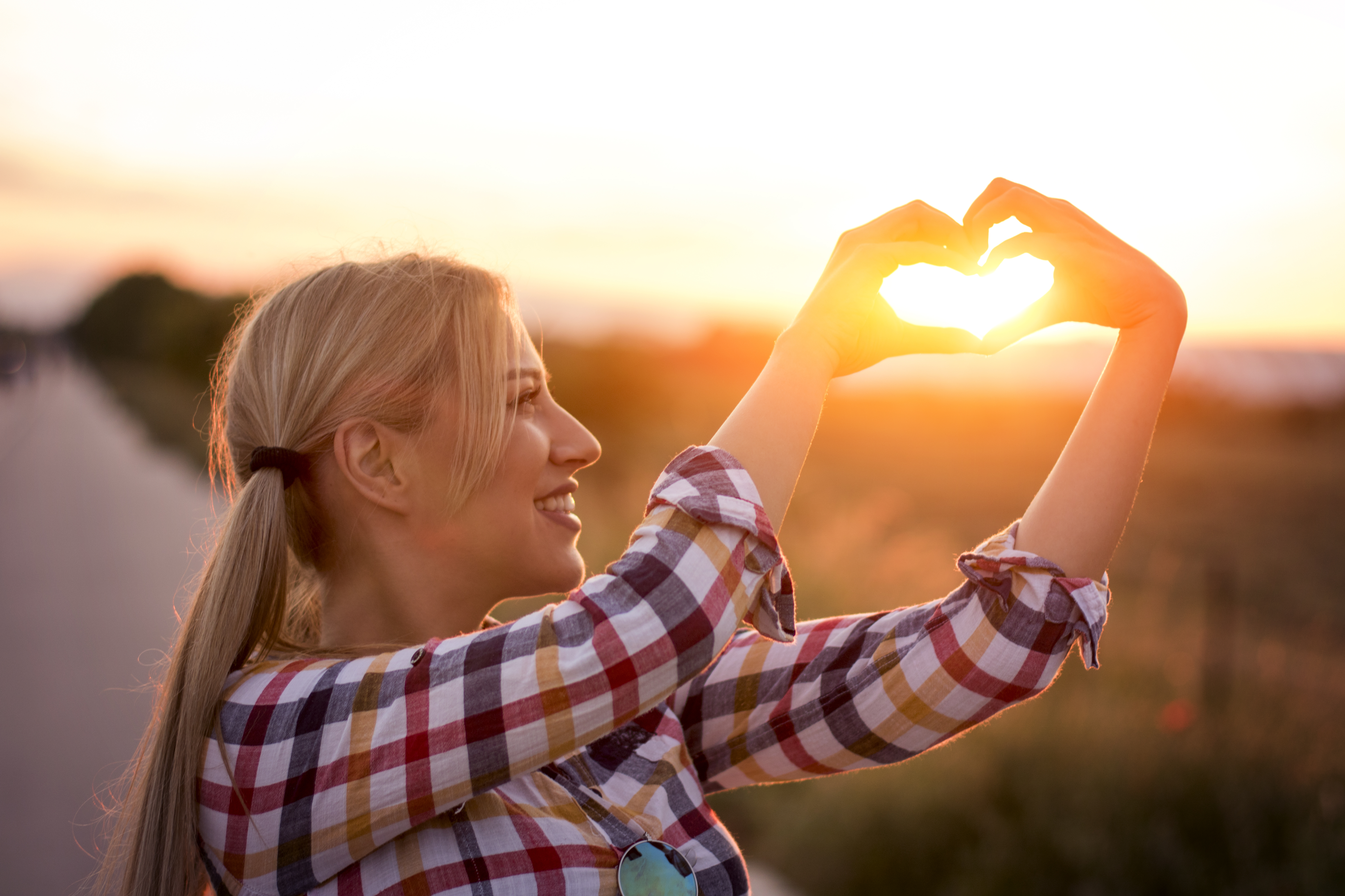 mujer con los dedos en forma de corazón y por en medio atraviesa una luz dorada del sol, paisaje de primavera